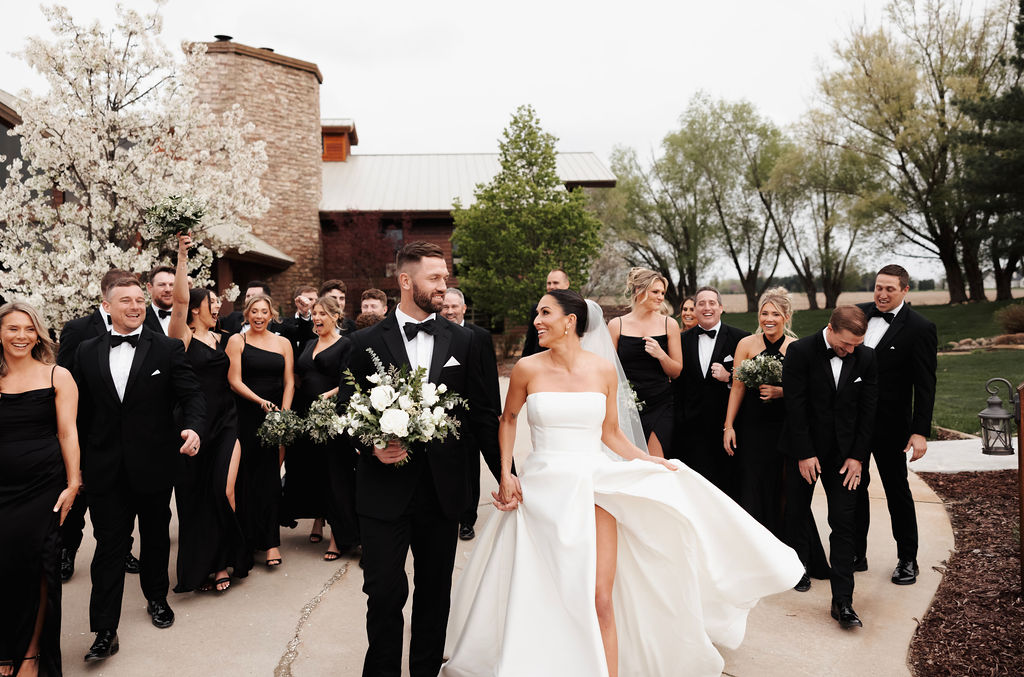 Bride and groom walking with their wedding party in black attire, carrying white and green floral bouquets.