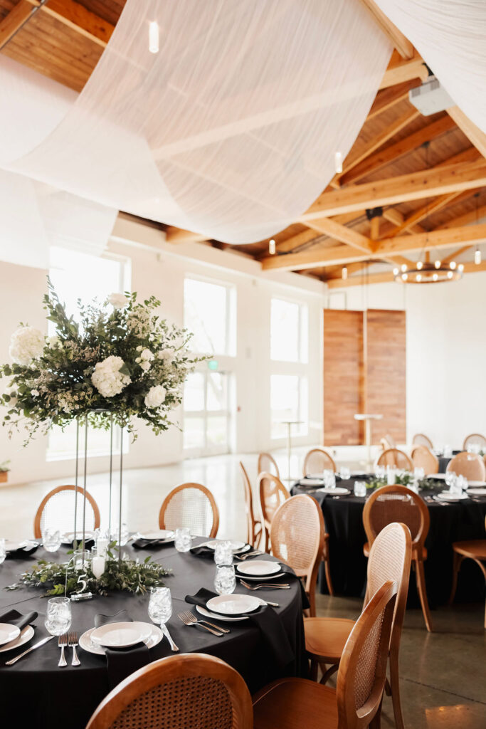 Wedding reception with black linens, wood chairs and tall white and green floral centerpieces.