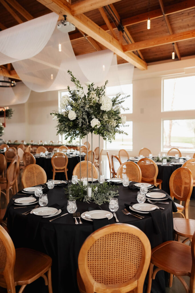 Reception table with black linens and a tall white floral centerpiece surrounded by greenery and candles.
