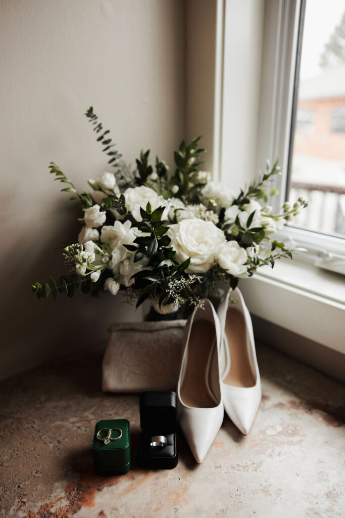 White and green bridal bouquet displayed beside white wedding shoes and wedding rings near a window.