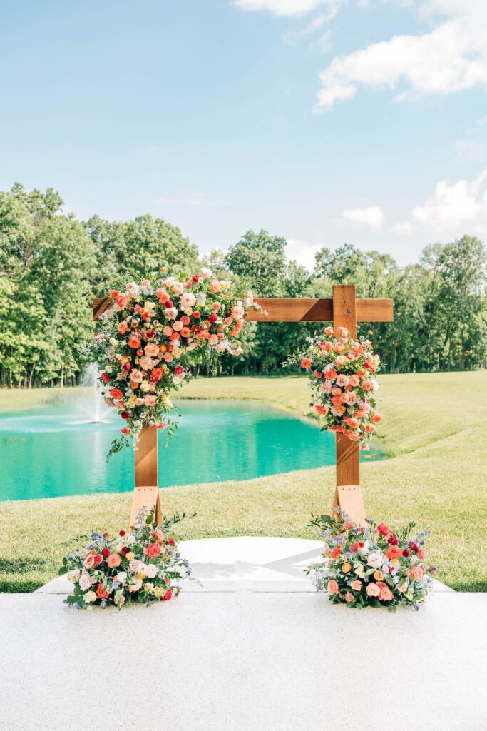 Outdoor wedding ceremony arch covered in colorful coral, pink and peach flowers beside a pond.