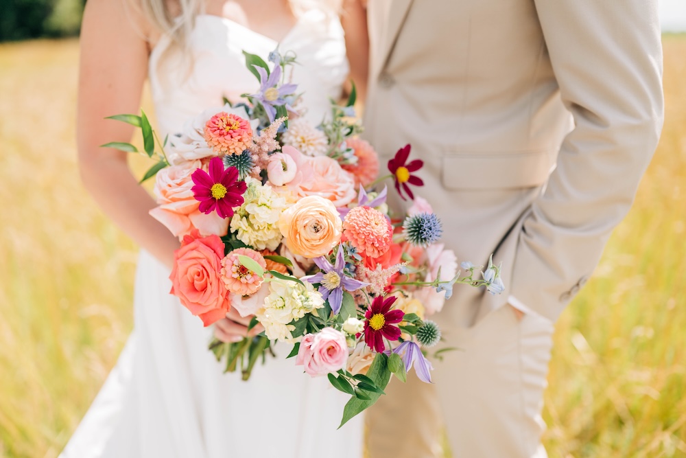 Bride and groom holding a colorful wedding bouquet with coral, peach, pink and blue flowers.