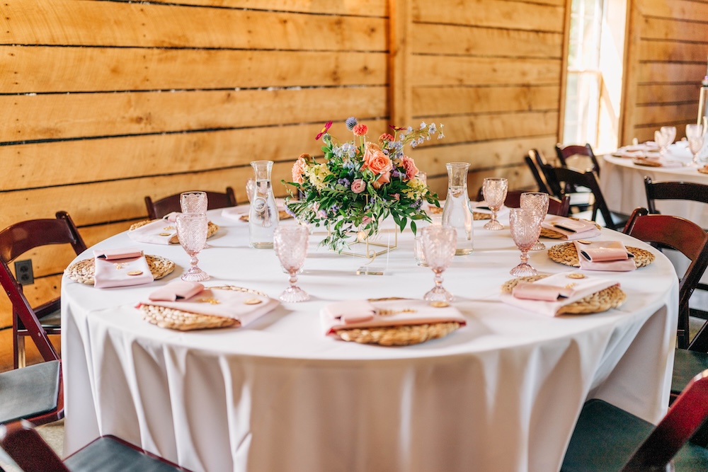 Colorful garden-style floral centerpiece on a reception table with blush glassware and woven place settings.