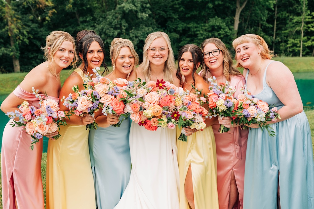 Bride and bridesmaids holding colorful garden-style bouquets in shades of peach, pink, yellow, blue and lavender at an outdoor summer wedding.
