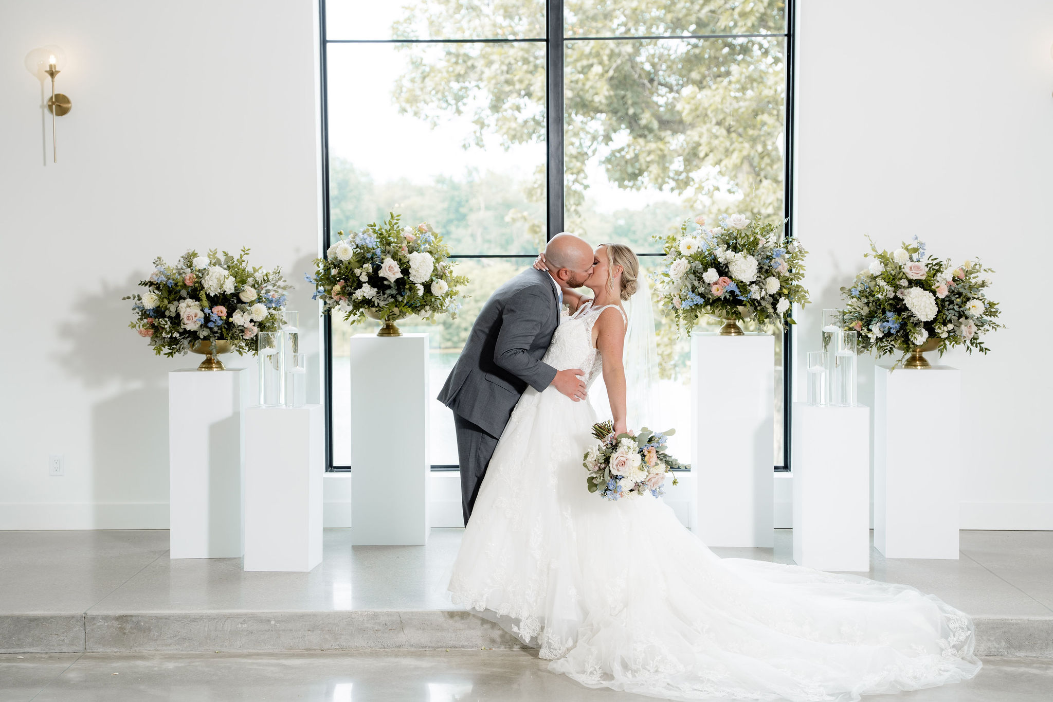 Bride and groom kissing in front of pastel wedding floral arrangements displayed on white pedestals.