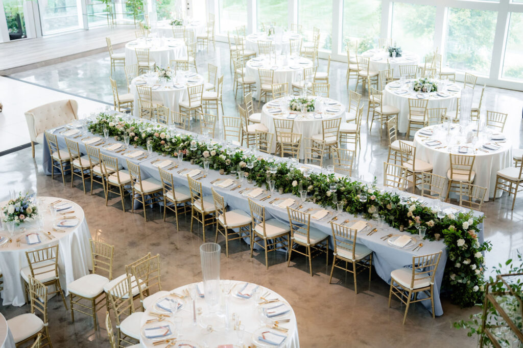 Wedding reception with a long head table lined with lush greenery and pastel flowers surrounded by round guest tables.