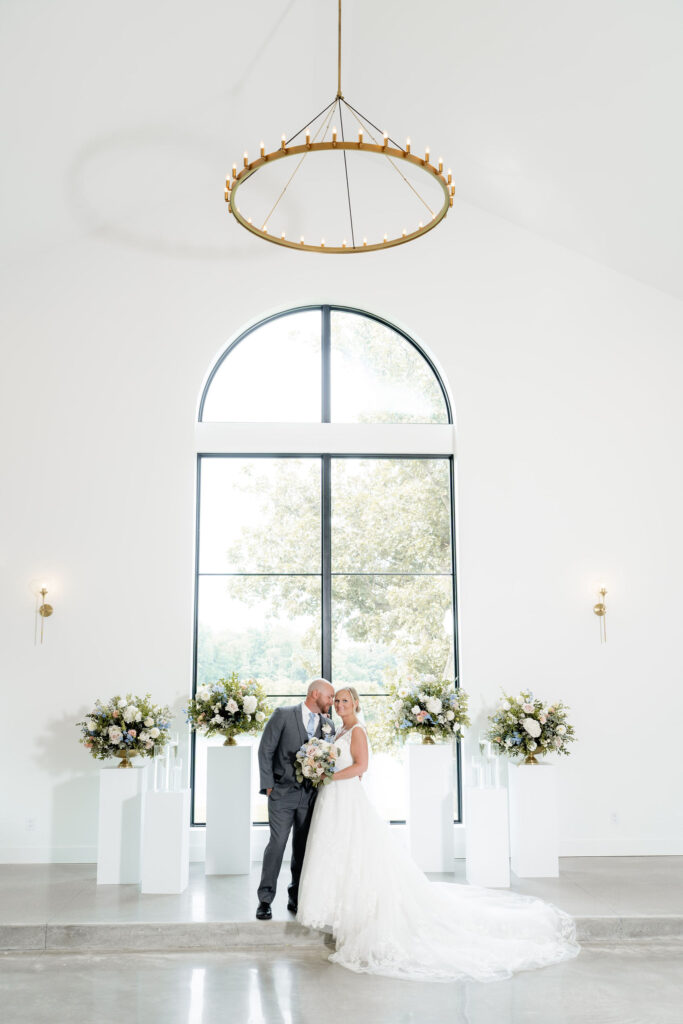 Bride and groom posing beneath a large arched window surrounded by pastel floral arrangements on white pedestals.