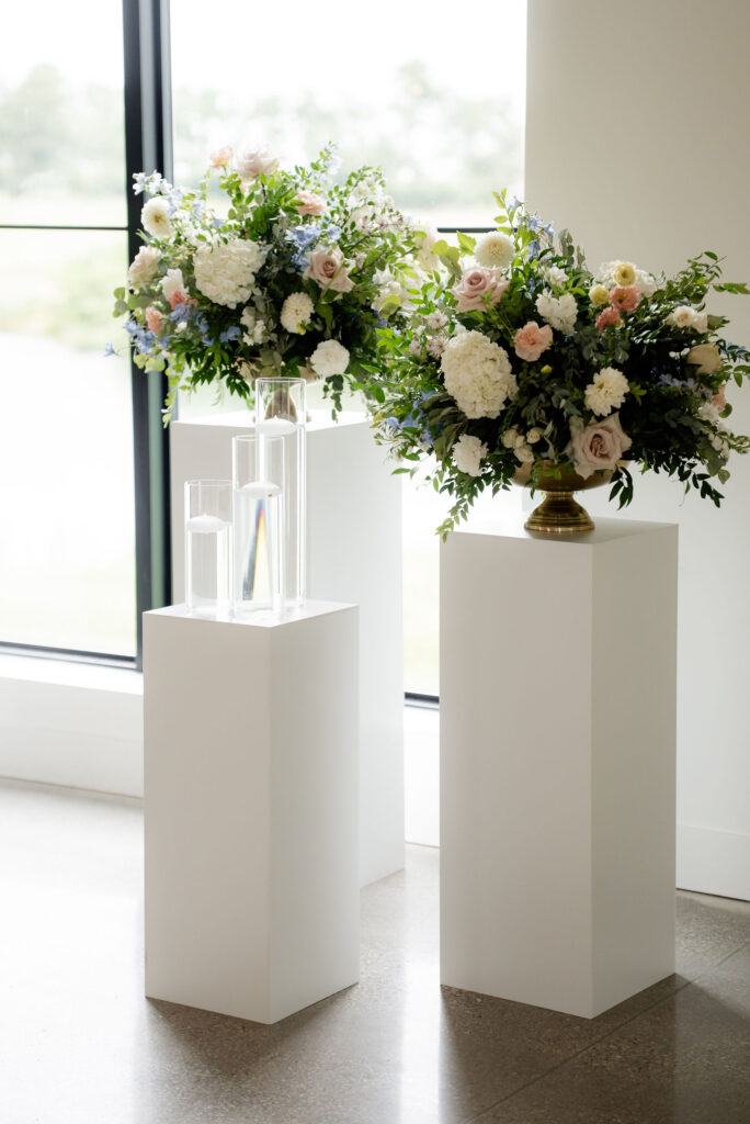 Pastel wedding floral arrangement with white, blush and blue flowers displayed on a white pedestal.