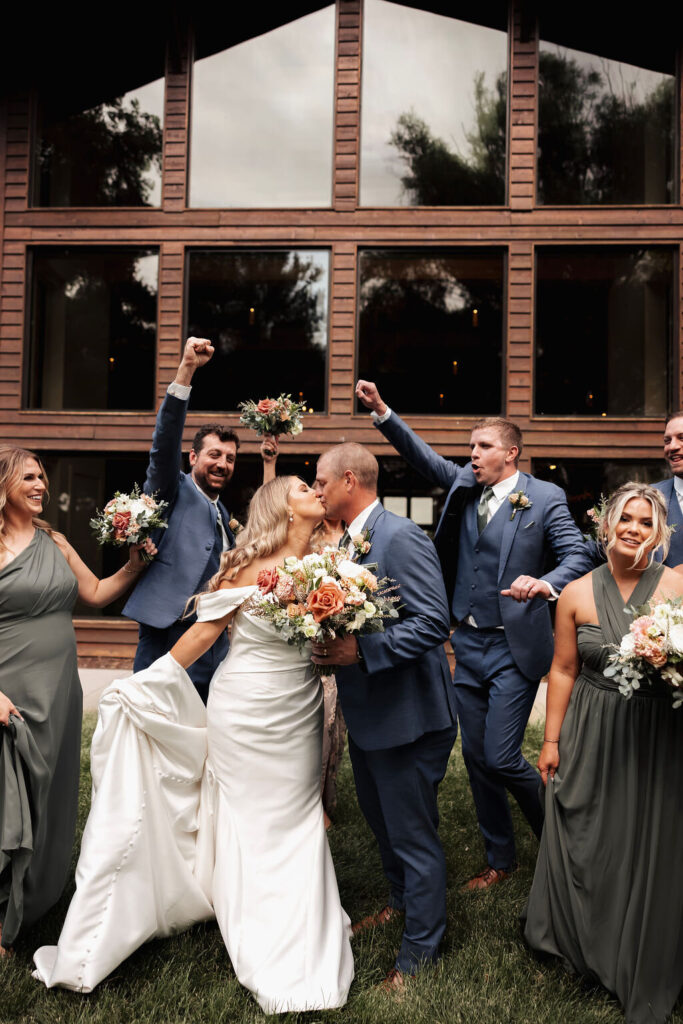 Bride and groom kissing with their wedding party outside a rustic venue, holding colorful floral bouquets.