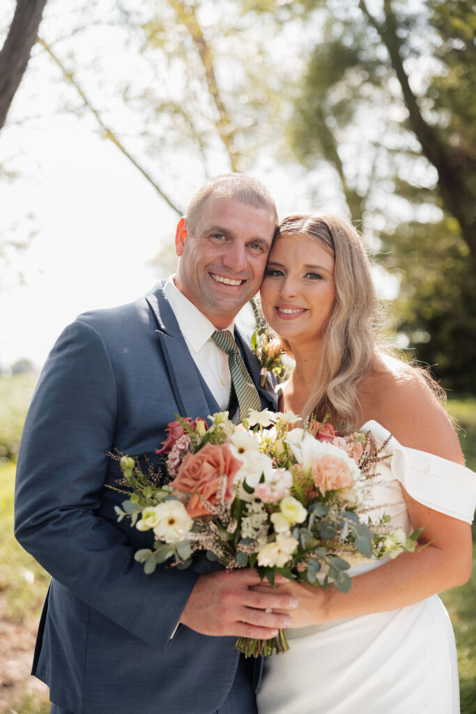 Bride and groom posing outdoors with a bouquet of peach, white and blush flowers.
