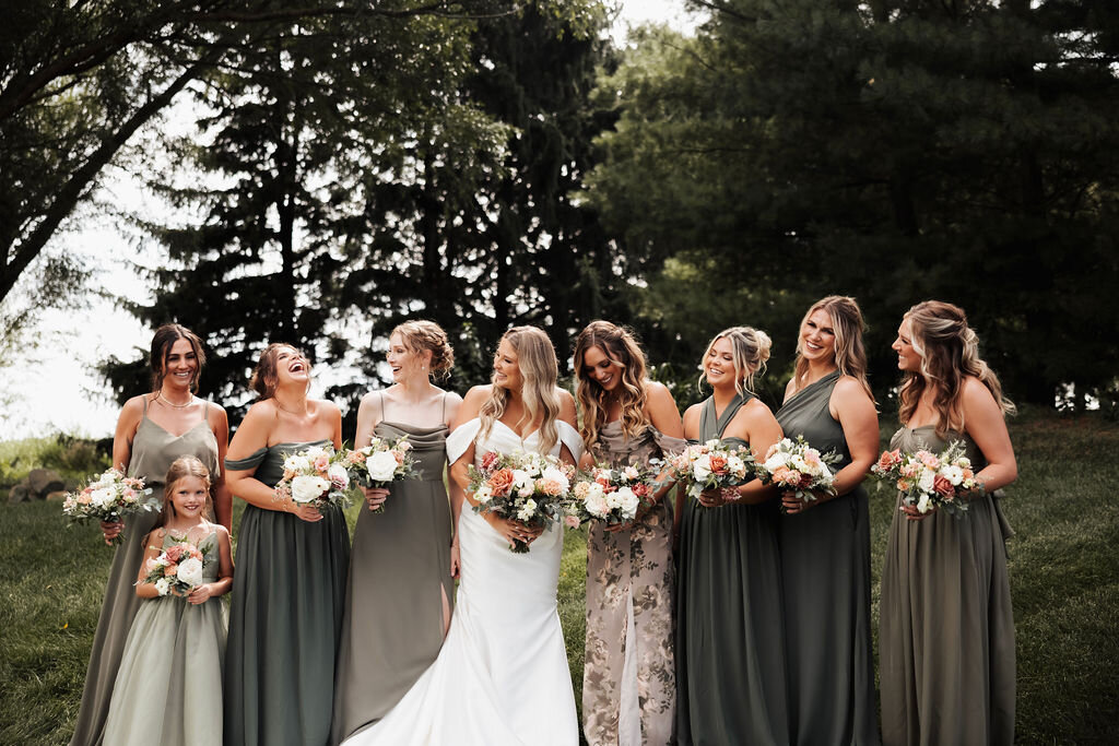 Bride and bridesmaids outdoors holding coordinating bouquets of peach, blush and white flowers with greenery.