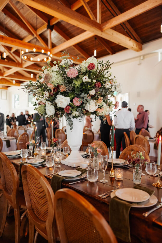 Tall wedding centerpiece with peach, blush and white flowers and greenery on a wooden reception table.