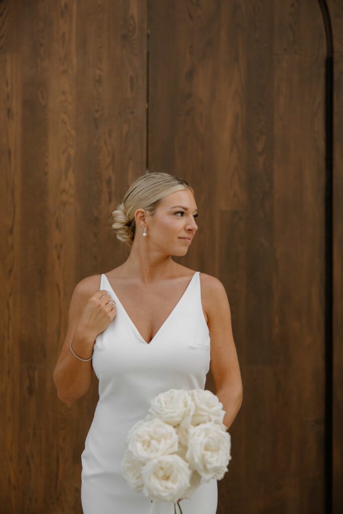 Bride in a white gown holding a bouquet of white roses against a warm wood backdrop.