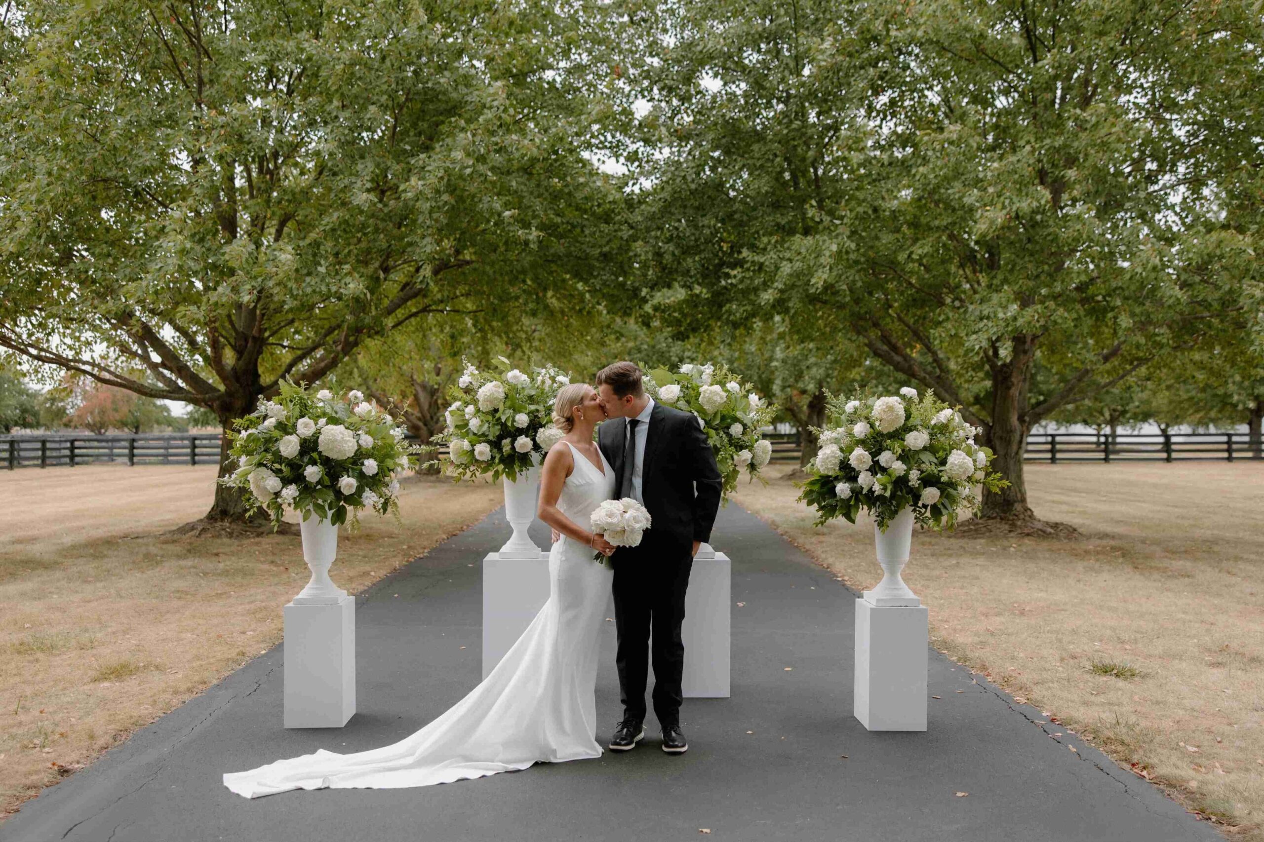 Bride and groom kissing beneath mature trees, surrounded by large white and green floral arrangements on white pedestals.