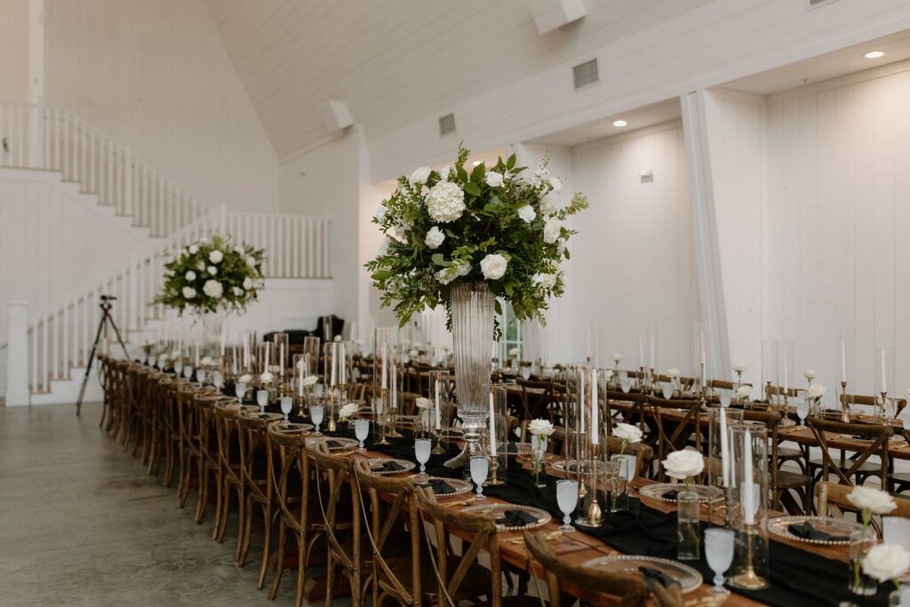 Wedding reception with long wooden tables, tall white and green floral centerpieces, black linens and candlelight.