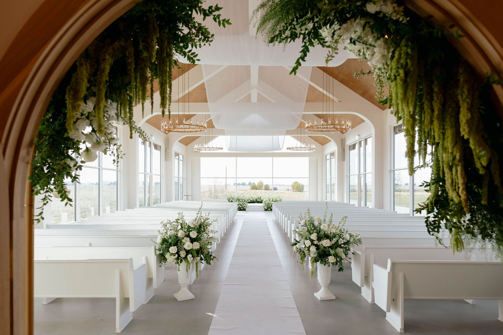 White wedding ceremony aisle at Pear Tree Estate framed by lush greenery and white floral arrangements.