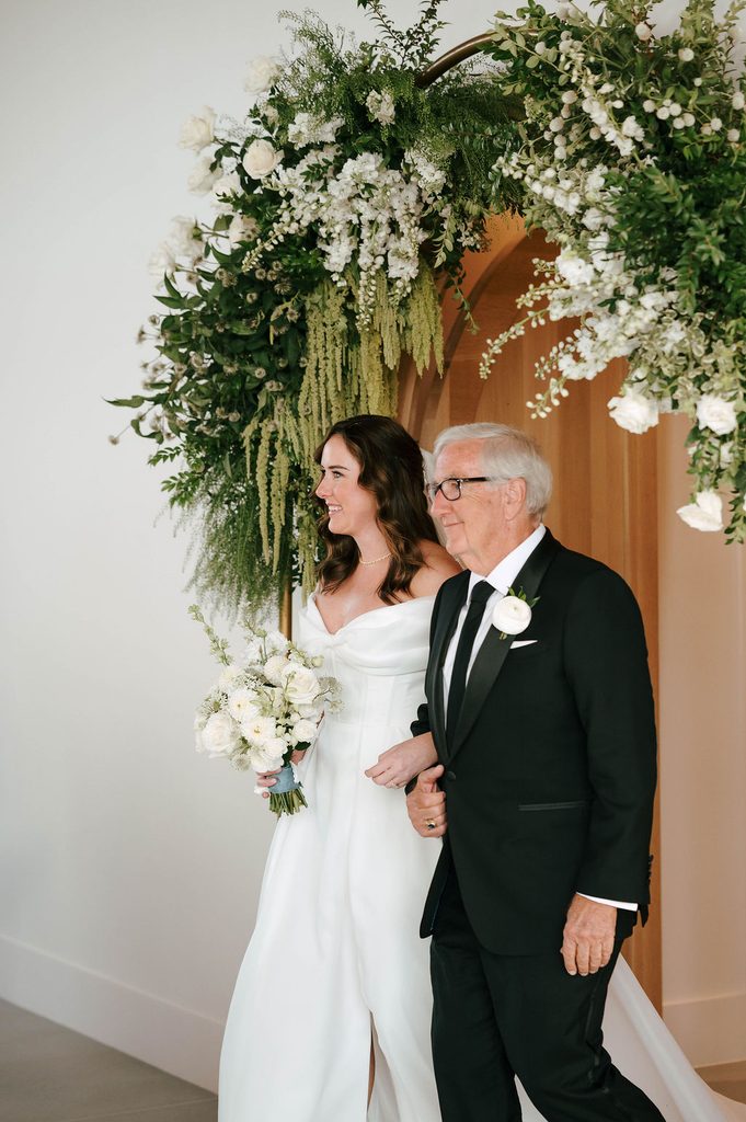 Bride entering the wedding ceremony with her father beneath a lush white and green floral arch at Pear Tree Estate.
